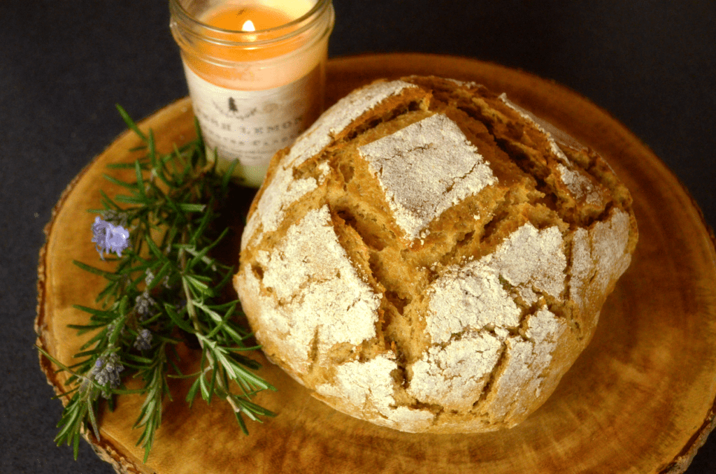 Wholegrain boule bread with rosemary sprig and lit candle on a round rustic wood cutting board.
