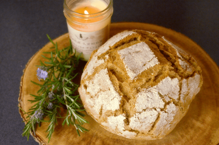 wholegrain einkorn boule bread sits on a rustic wooden cutting board nest to sprig of blooming rosemary and lit candle in mason jar.
