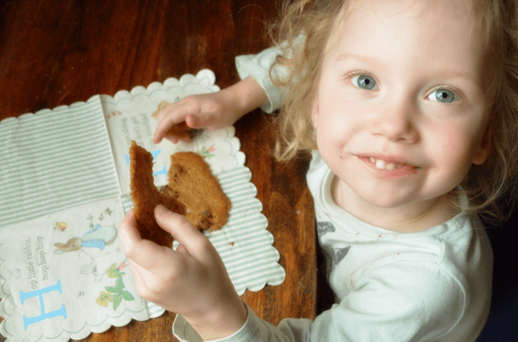 Little blue eyed girl smiles at camera while easting a chocolate chip cookie.