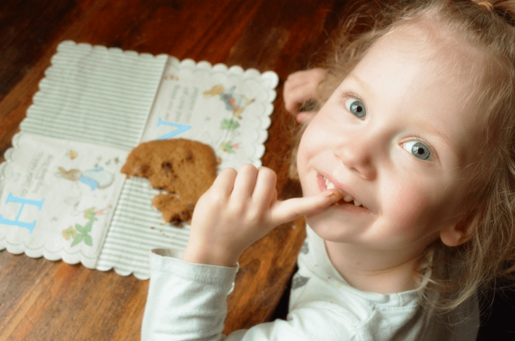 Camera looks down at blue eyed girl who smiles back with a chocolate stained finger touching her lips. A chocolate cookie sits half eaten at the table in from of her on top of a pastel colored napkin with peter rabbit.