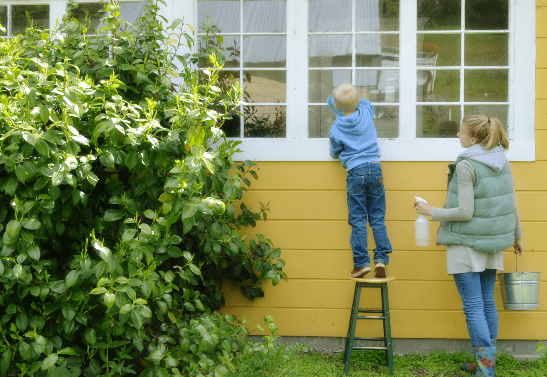 child stands on stool cleaning window while mom stands by and watches
