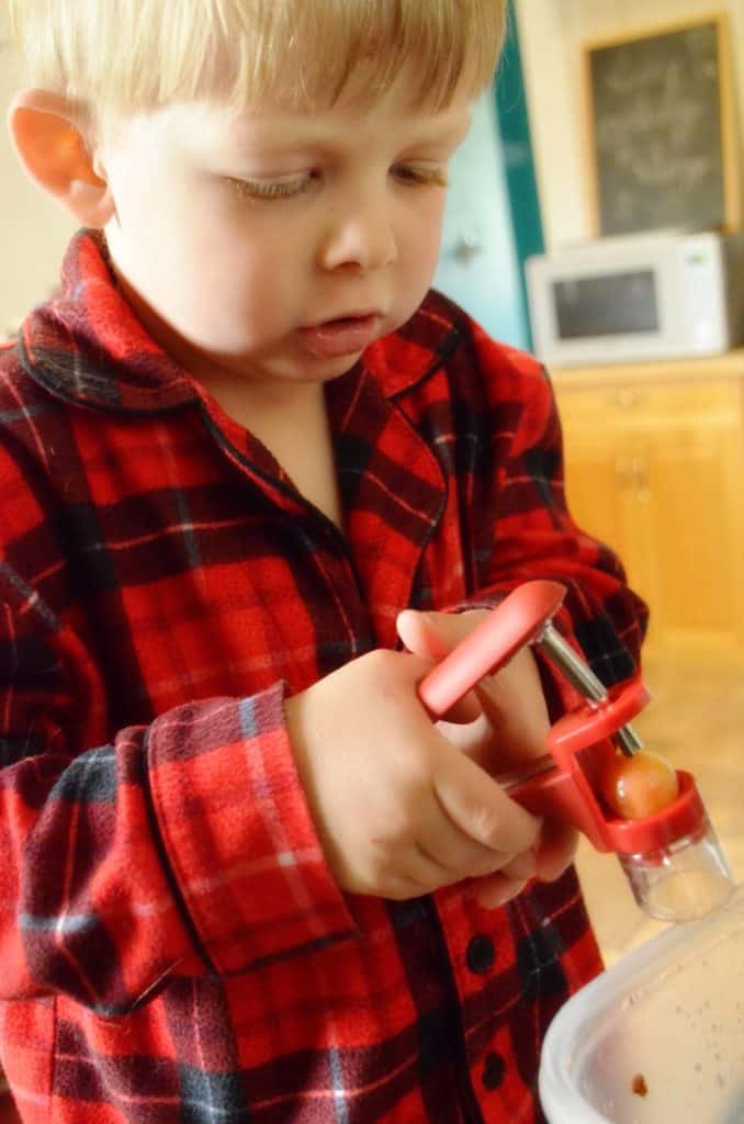 A toddler dressed in red plaid pajama's focuses on his job at hand of pushing a cherry through the pitter.