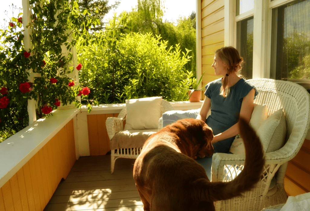A woman smiles sitting in a wicker loveseat on a porch. the evening sun streams in and she looks faraway past the dark pink roses climbing up a trellis along the porch edge. a brown Labrador pokes his head in her lap looking for attention .
