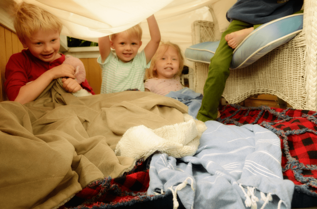 Children play under blankets and sheets inside a home made fort. A large sheet is held up over them like a tent by the wicker seating on a porch.