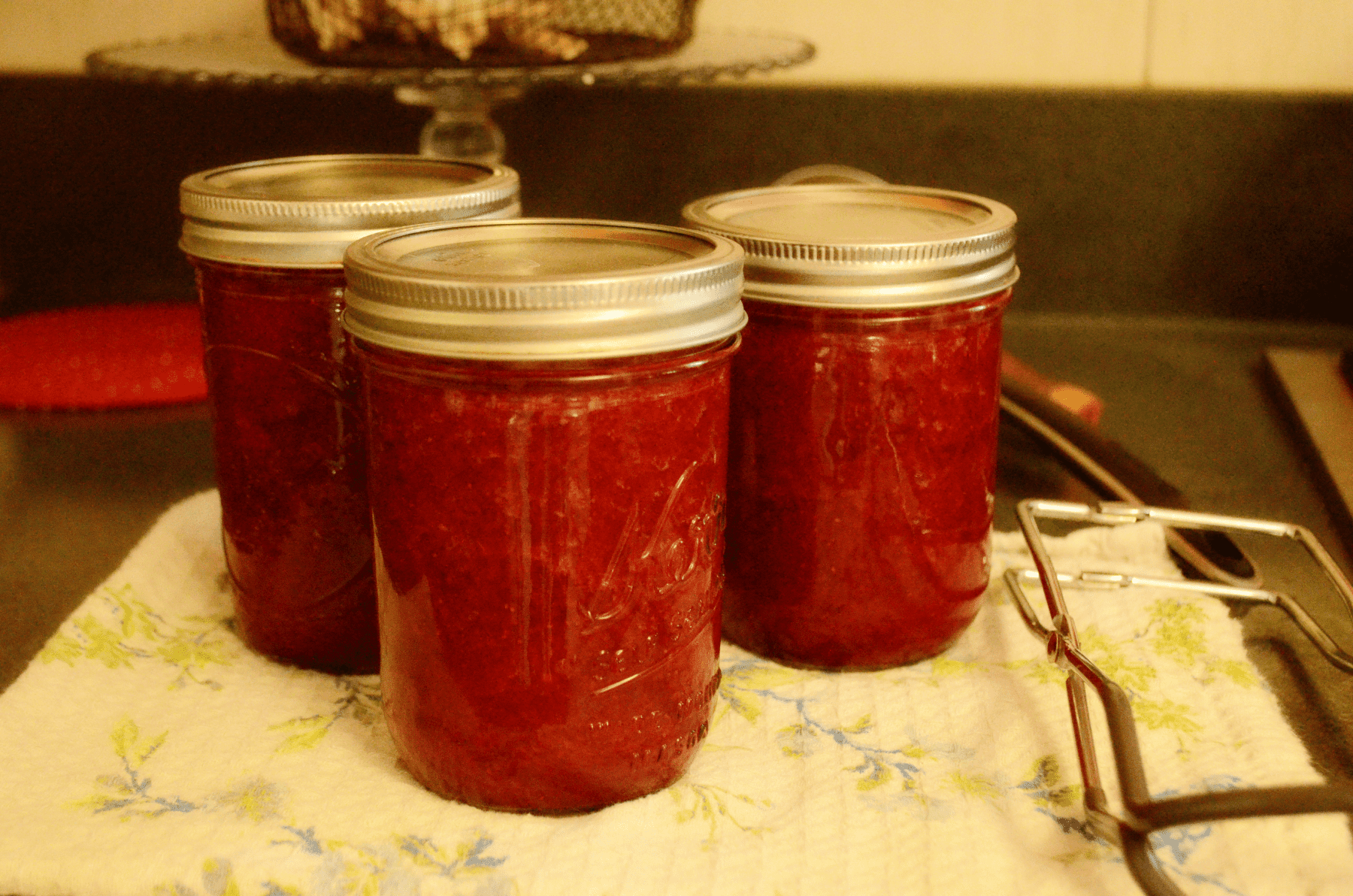 Three jars are filled with fresh strawberry jam cooling on a floral towel. Next to the jars lies a canning jar lifter.