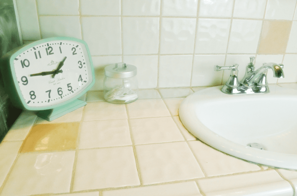 A mint colored clock sits next to a glass container on a clean bathroom sink with a shiny cleaned chrome sink.