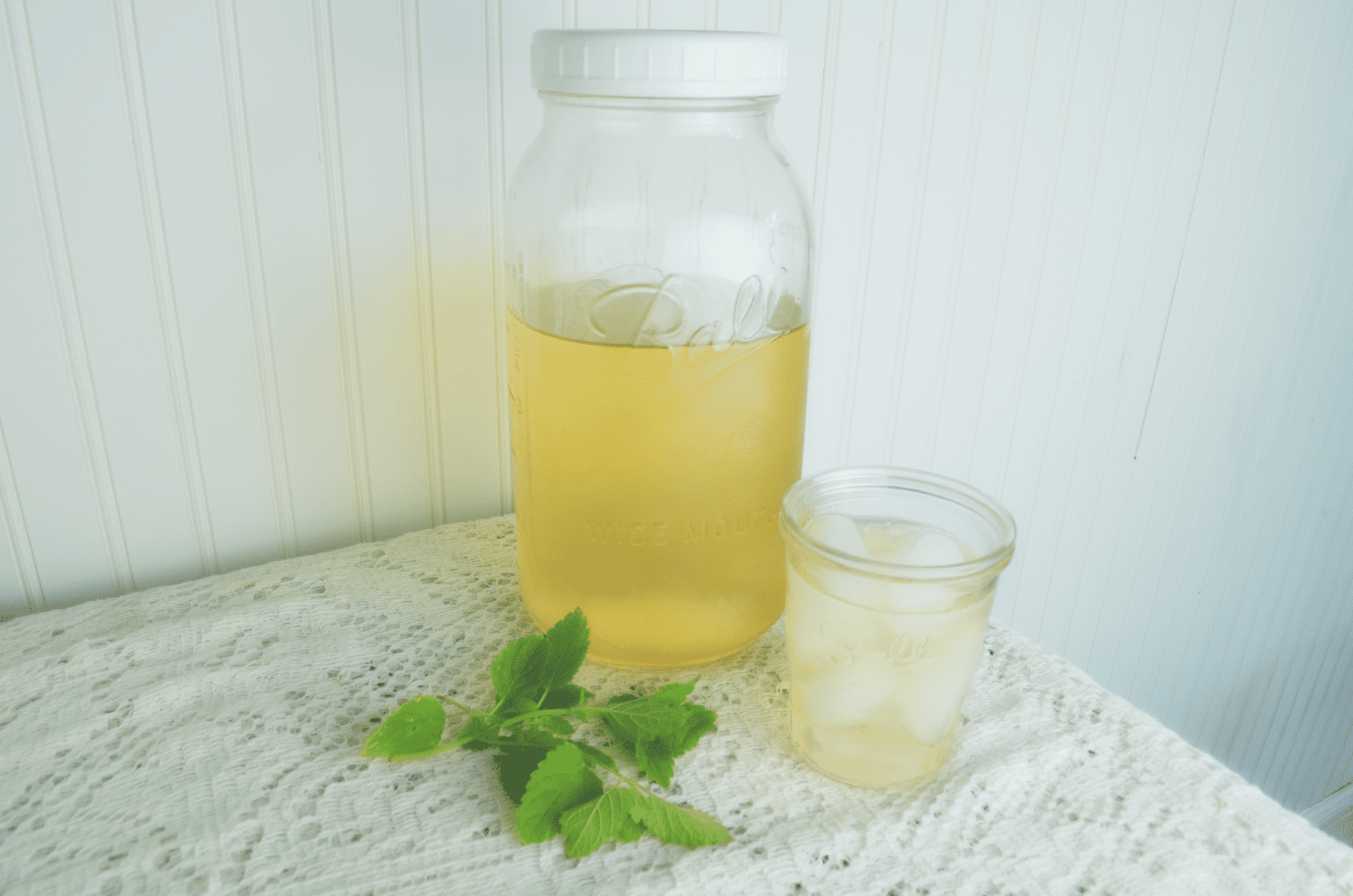 A large Ball canning jar filled with fresh iced tea sits on a lace tablecloth next to a cup of iced tea and green lemon balm leaves