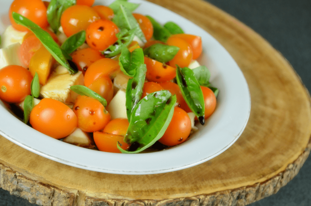 Juicy red cherry tomatoes, white chunks of cheese and fresh basil leaves a drenched with droplets of oil and balsamic vinegar in a small serving bowl on a rustic cutting board.