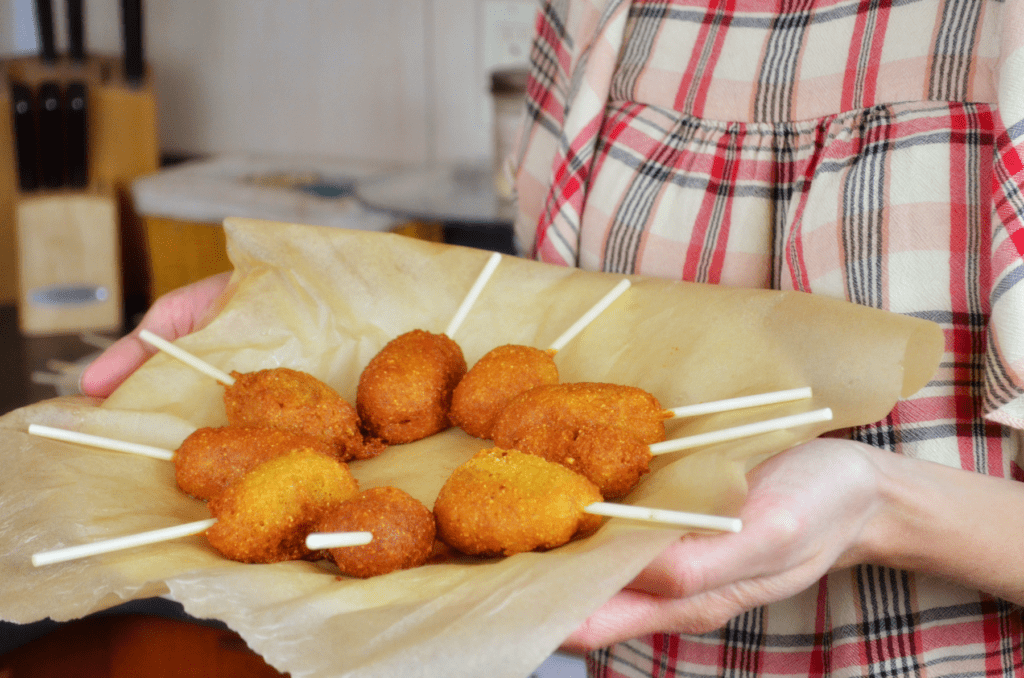 A woman in red and brown plaid carries in a tray of freshly fried corndogs.