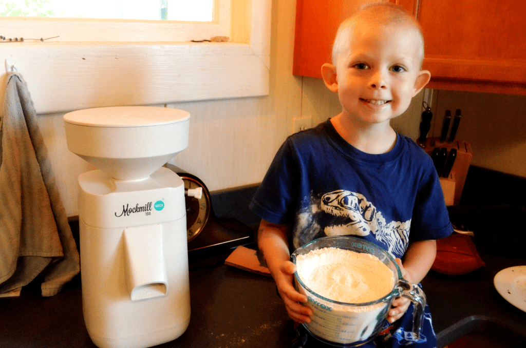 Little boy smiles  big holding a bowl of freshly ground flour while sitting on a countertop next to the Mockmill 100 flour mill 