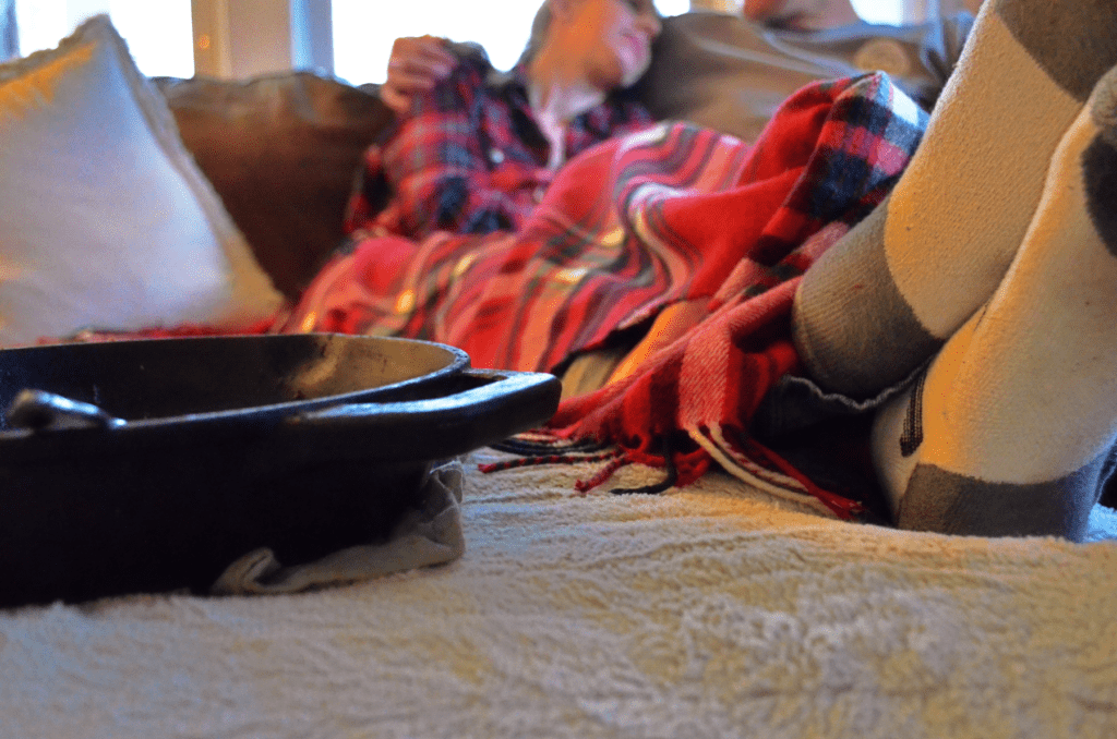 An Iron skillet sits in front of a couple cuddling on a couch. Feet are put up and a plaid cozy blanket keeps them warm.