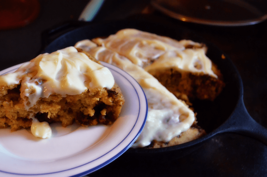 A Soft Einkorn Pumpkin Streusel Scones covered in gooey cream cheese frosting is lifted on a plate from an iron pan holding the rest