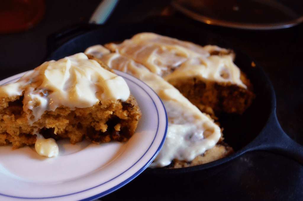 A plate filled with a soft frosting covered scone is held in front of an iron pan. The iron pan has just one piece cut from the delectable white cream cheese covered scones.