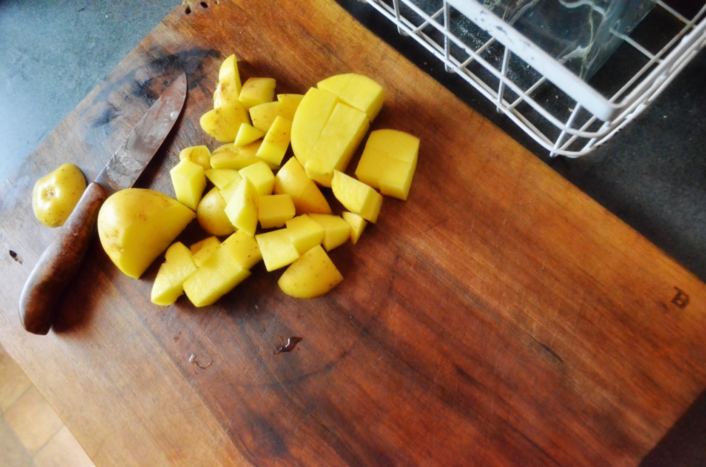 A Golden wooden cutting board shines from a recent oiling while a knife sits on top among freshly cut white potatoes.