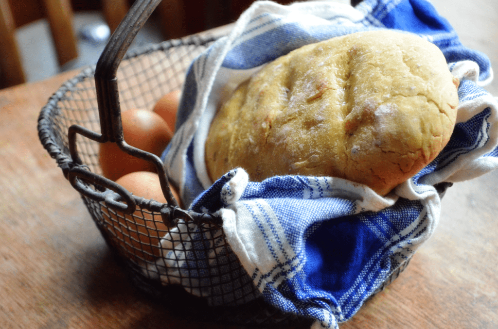 A wire basket is filled with fresh brown eggs sitting next to a towel wrapped loaf of freshly baked sourdough garlic herb bread.