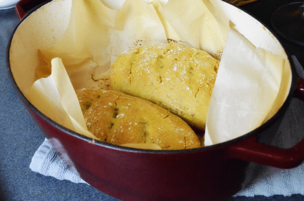 Two loaves of baked sourdough garlic herb bread sit side by side in a red dutch oven.