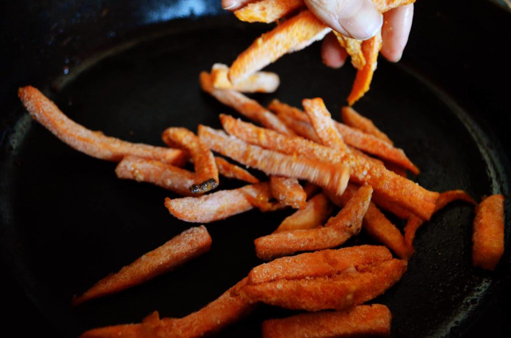 A hand drops frozen Alexia sweet potato fries onto an iron pan and spreads them out to get ready for oven baking.