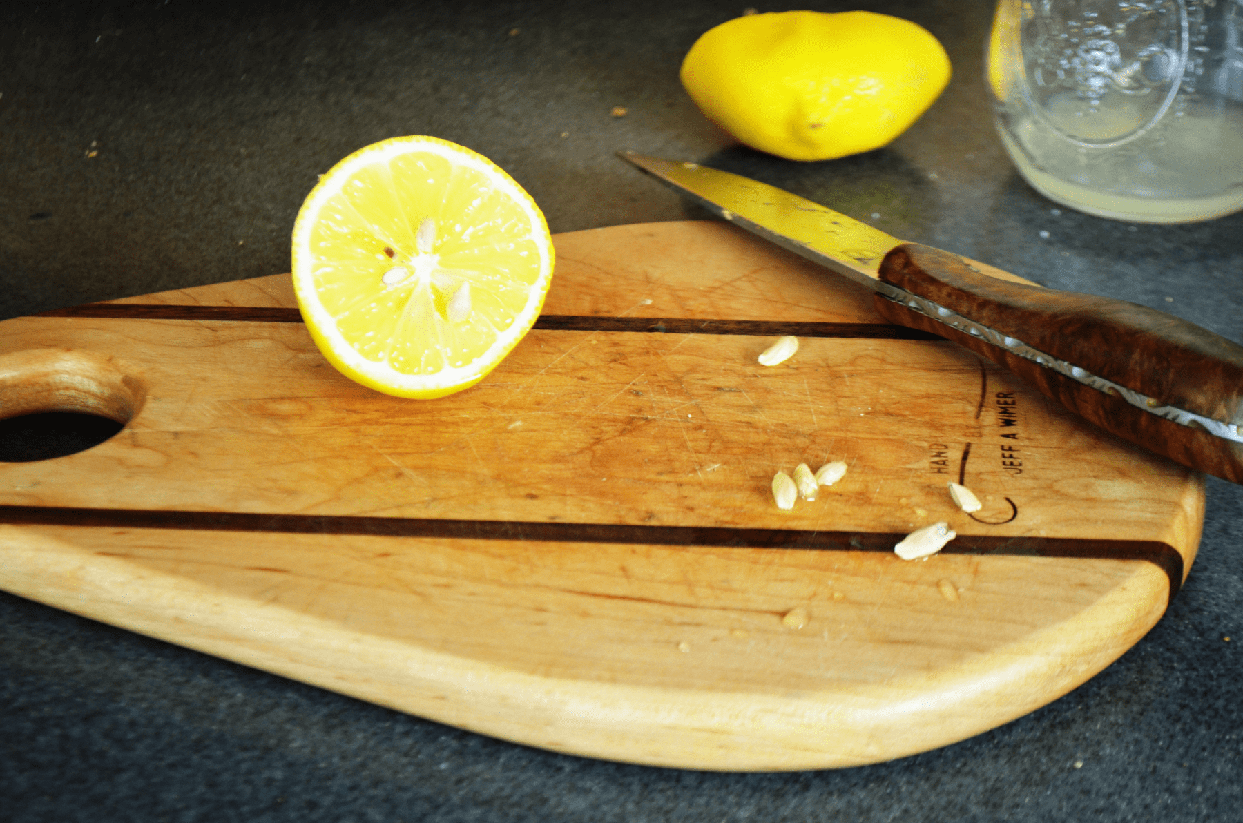 A-lemon-sliced-in-half-sits-with-a-wood-handled-knife-and-several-seeds-on-a-wooden-cutting-board.-Behind-the-board-sits-a-squished-squeezed-lemon-next-to-a-jar-with-lemon-juice.