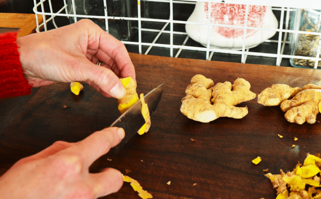 Hands reach into the image cutting peels from spicy ginger root. many yellow tinted peel scraps are seen to the side. Two large ginger fingers wait to be cut.