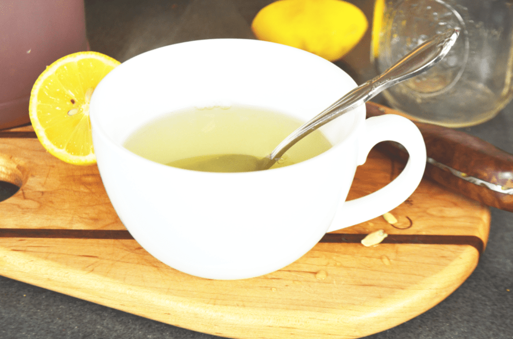 A chubby white bright up of ginger honey lemon tea sits atop a wooden cutting board. Two bright lemon halves lay behind it with a little glass jar of lemon juice.