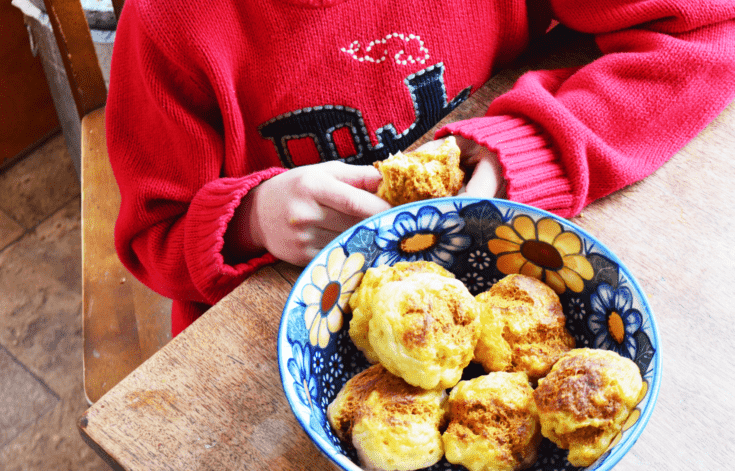 Gluten free rolls sit in a flowery blue bowl while a boy with a red sweater sits in frnt of the bowl munching on a half eaten roll.