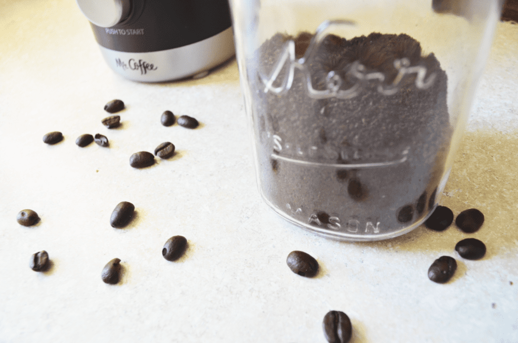 A Mason Jar holds ground coffee while coffee beans lay scattered around it on the countertop. 