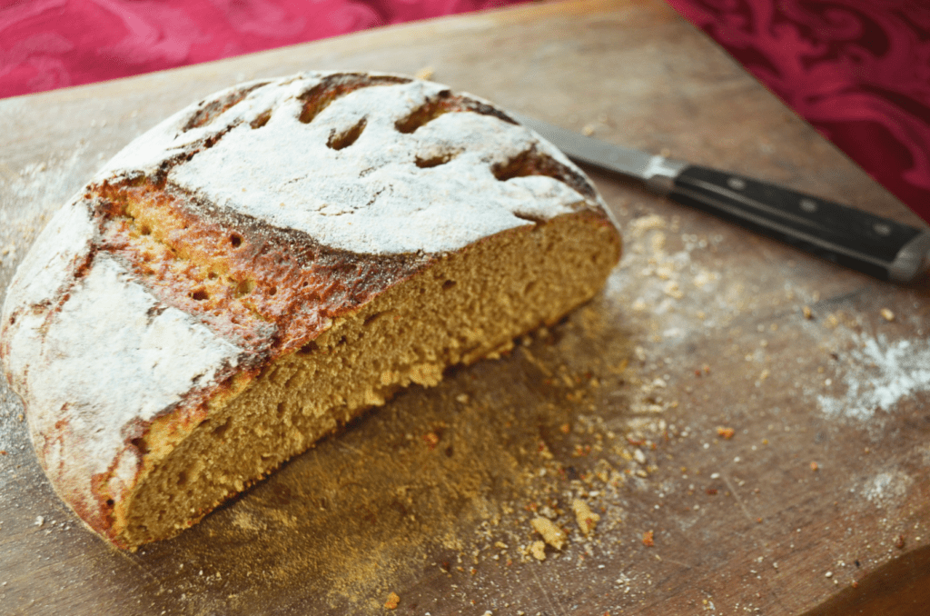 A whole grain artisan style round bread sits with a white flour covered top but for the cracks where it rose in the oven. It is sliced open with crumbs and flour covering the wooden cutting board where it stays. A shiny knife lies to the back offering assistance to any who want a slice of fresh bread. This is one of the best breads for acid reflux.