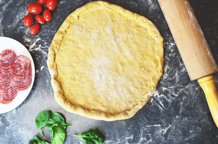 A buttery colored dough rolled into a circle sits atop a floured countertop with bright red tomatoes, meats and green spinach to top the pizza.