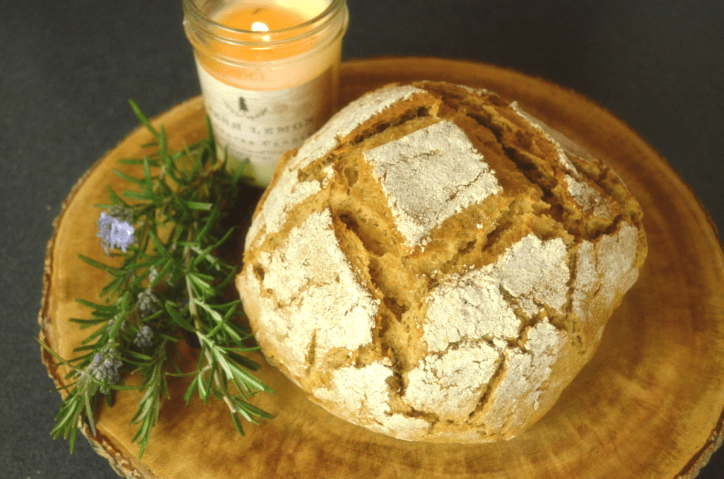 A whole wheat artisan loaf shares a round wooden cutting board with a small mason jar candle and fresh sprigs of rosemary herbs with lavender flowers budding.