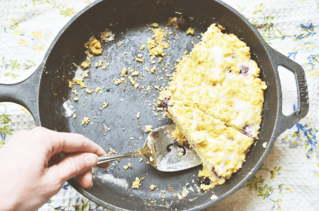 A hand reaches to scoop a slice of icing drizzled lemon blueberry einkorn scones out of a dark iron pan.