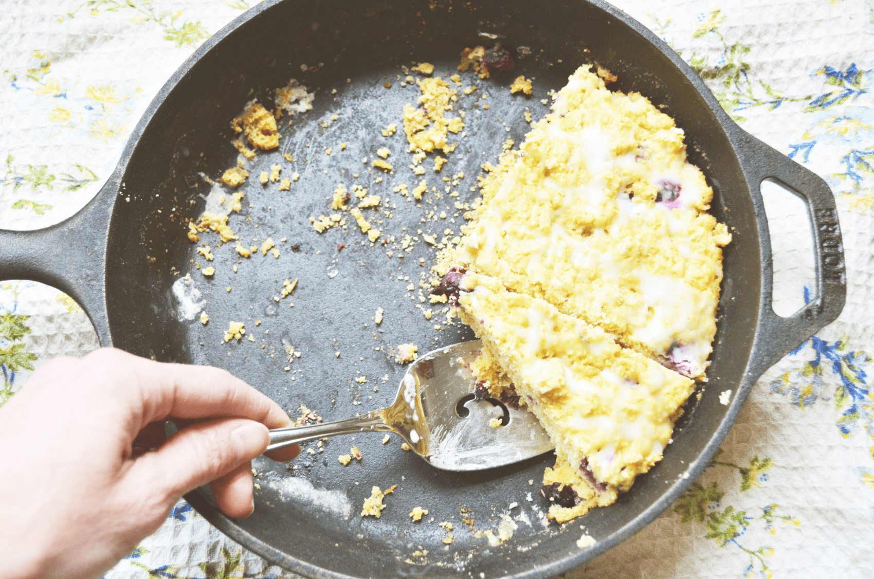 a hand reaches to scoop out a bright icing drizzled slice of blueberry lemon einkorn flour scones in an iron pan.