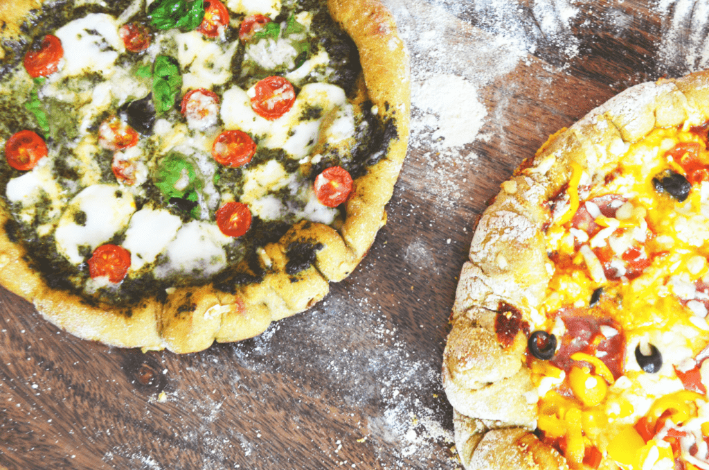 Two thick crust pizzas sit on a floured wood cutting board. Their toppings still bubble and glisten from being just taken from the oven. Their crusts are a crunchy brown.
