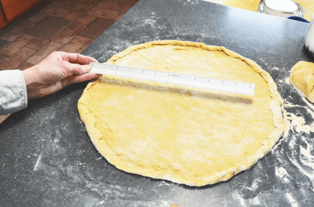 a large rolled out einkorn pizza dough lies on a floured countertop. A hand holds a ruler above the dough to measure it's diameter.