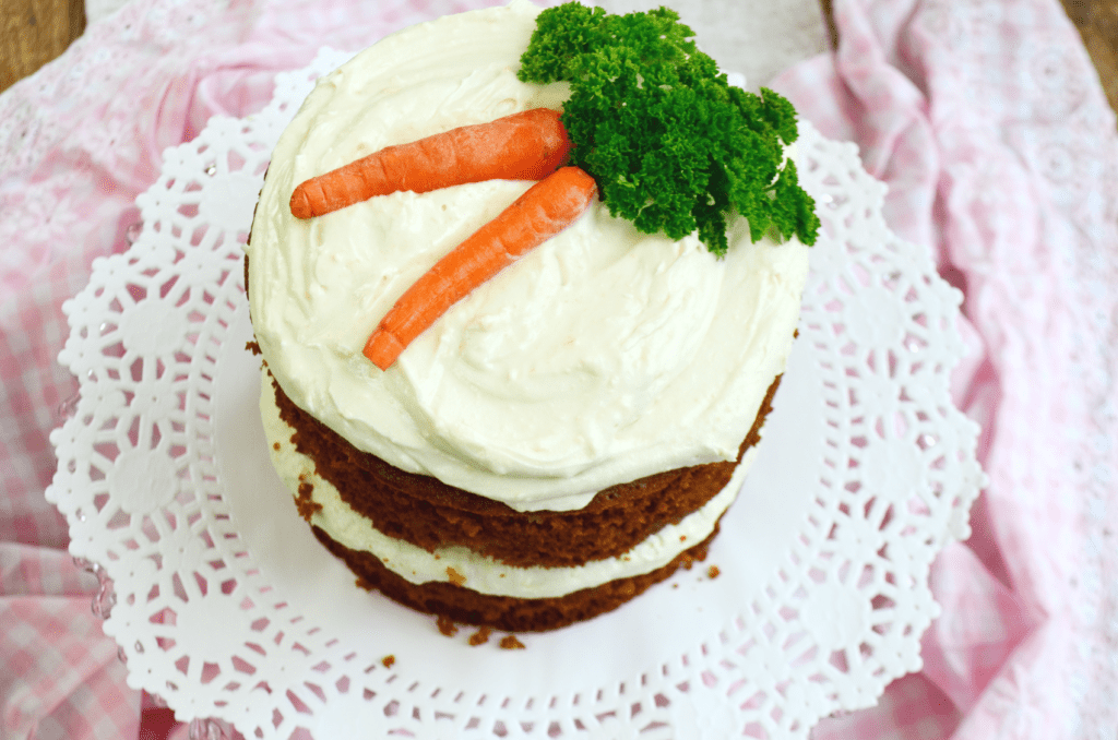 Two mini carved carrots with parsley for the greens top a tall round dark carrot cake with bright white frosting.