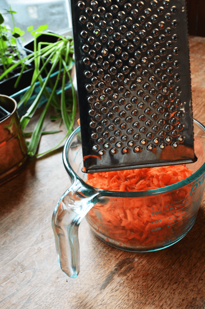 Bright orange carrot bits a re grated by a shiny silver grater into a large glass measuring cup