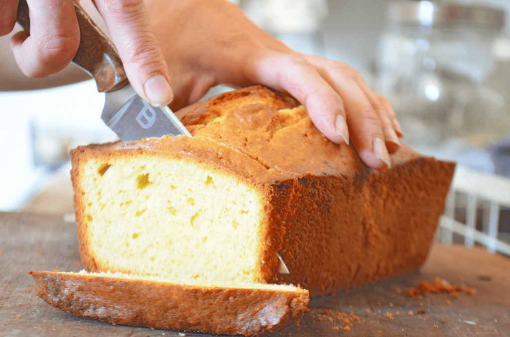 Hand drives a knife through a browned loaf of sweet lemon pound cake, cutting slices.