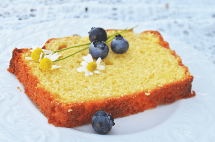 A slice of rich lemon pound cake lies with blueberries and flowers on a white plate.