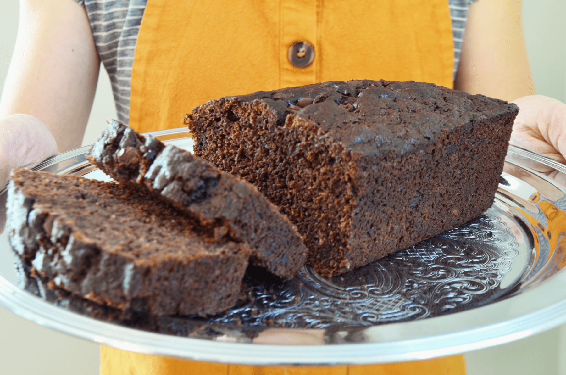 hands hold a silver platter carrying soft dark chocolate bread with bits of chocolate chunks sprinkled throughout.