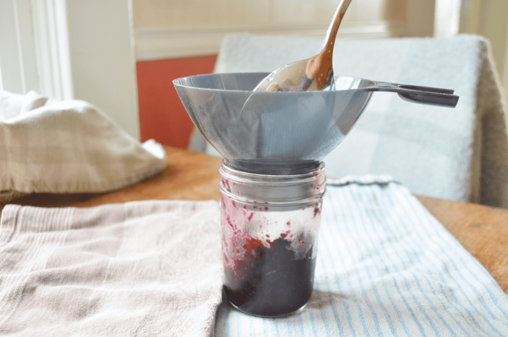 A shiny silver spoon fills blueberry preserves into a glass jar through a funnel.