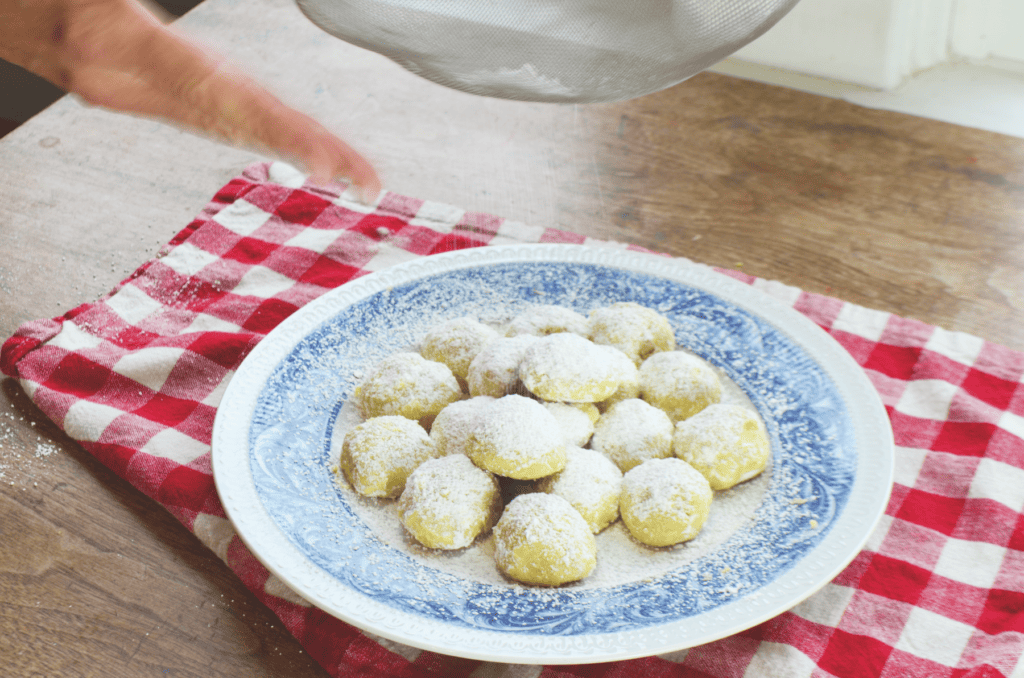 A hand gently beats a sifter shaking powdered sugar upon snowball cookies.