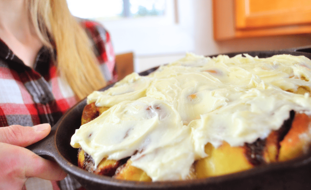 A Woman holds an dark iron pan filled to the brim with gooey cinnamon rolls topped with light sticky frosting.