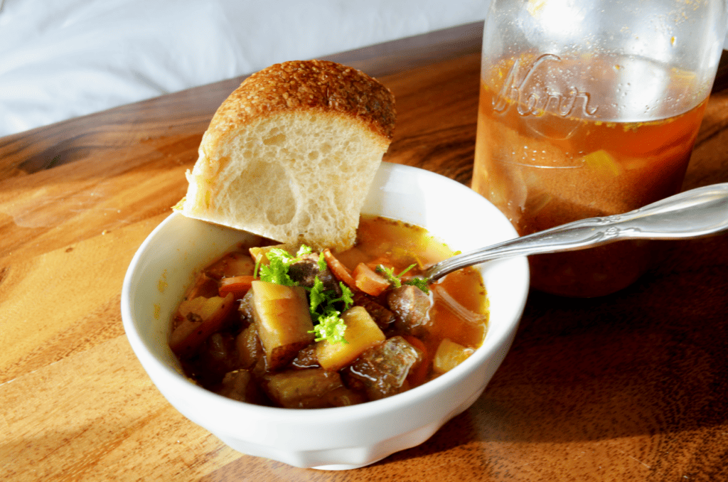 white bowl of hot beef stew garnished with bright fresh parsley sits on a smooth wooden table next to opened canning jar.