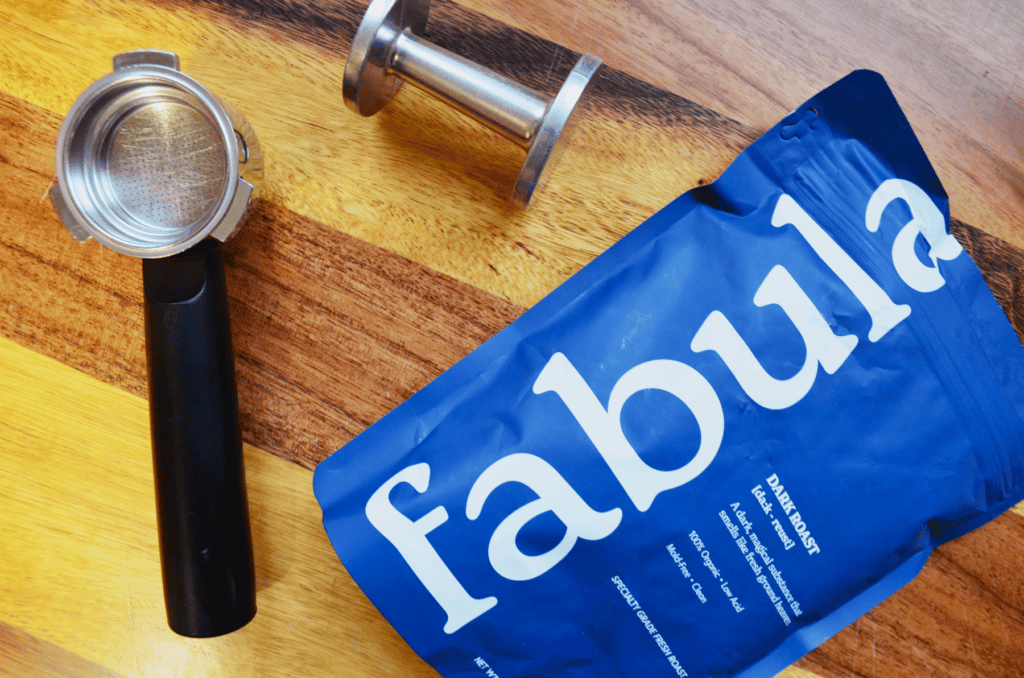 A blue bag of Fabula coffee sits on golden wooden table with a tamper and a portafilter