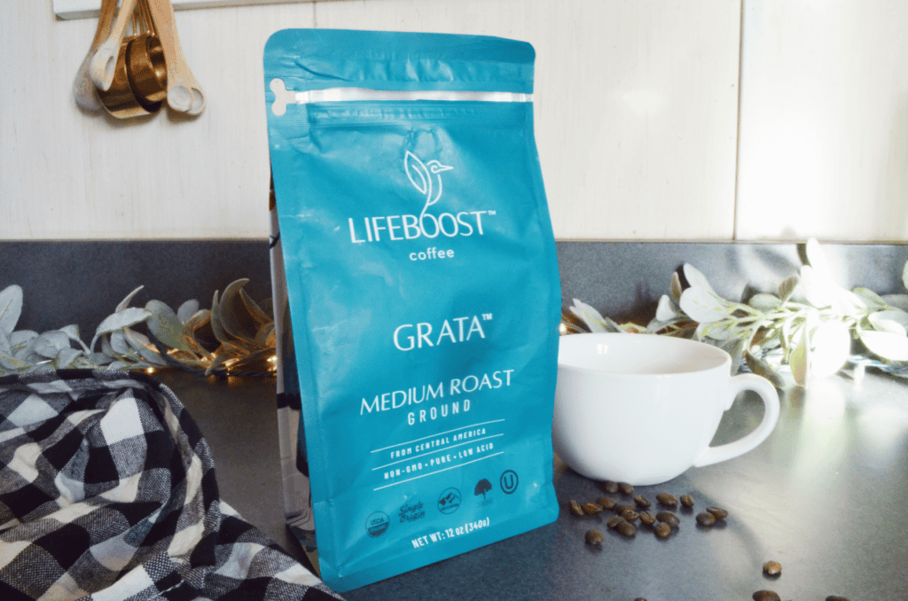 A Lifeboost Coffee Bag sits on a counter among roasted coffee beans and a qhite mug.